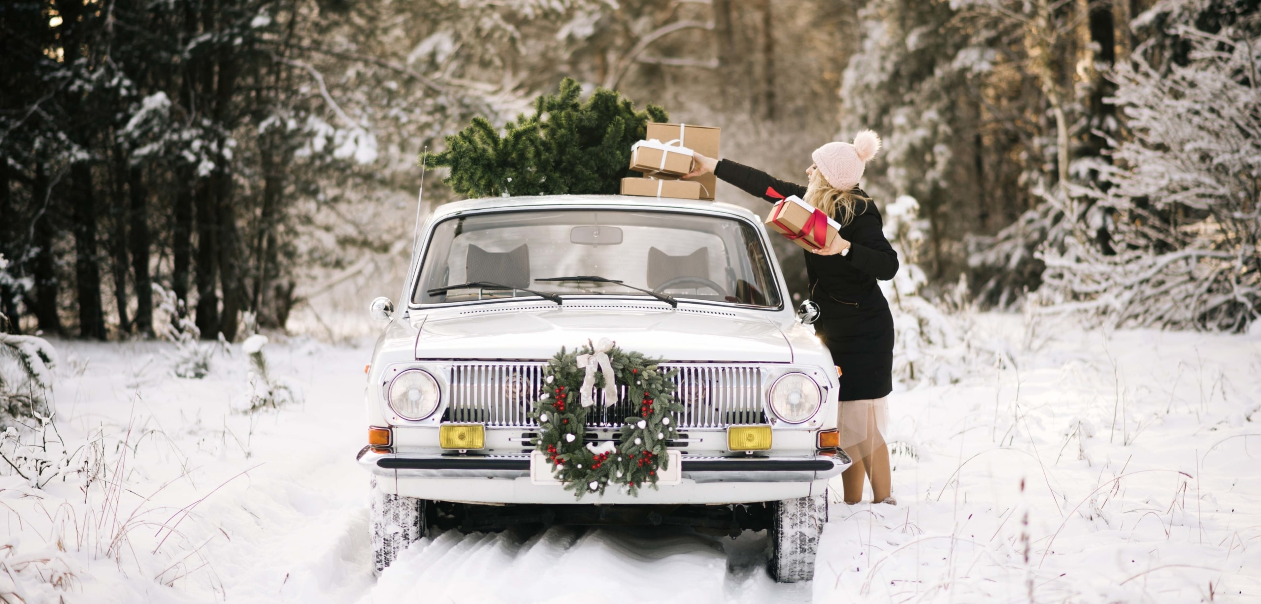 A vintage car with a decorative wreath on the front is parked in the snow as a woman in a winter hat piles Christmas presents on to the roof | Holiday Car Buying Could Mean Tree-mendous Savings