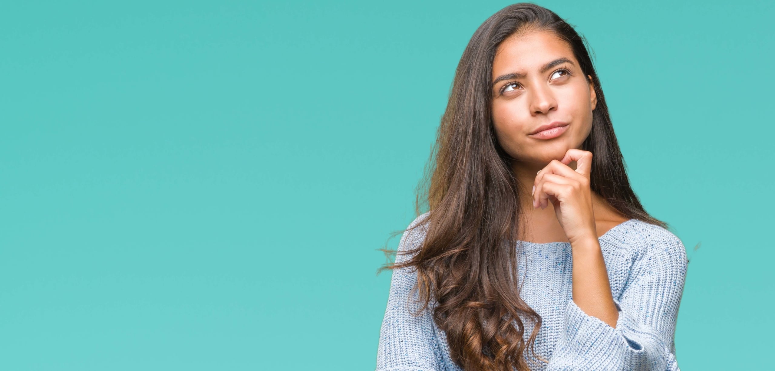 Young South Asian Woman with brown hair and a light blue knit sweater looks pensively into the upper right corner with her hand on her chin | 8 Car Insurance Questions To Refinance