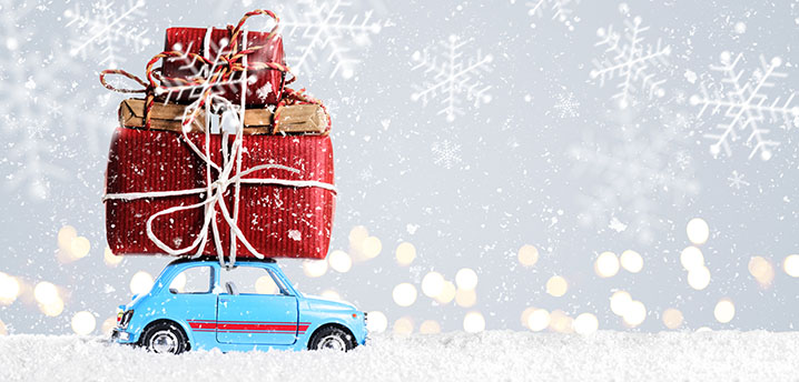 A blue car toy is driving through decorative snow with gifts piled up and tied to the roof | Gift Guide for Car Lovers