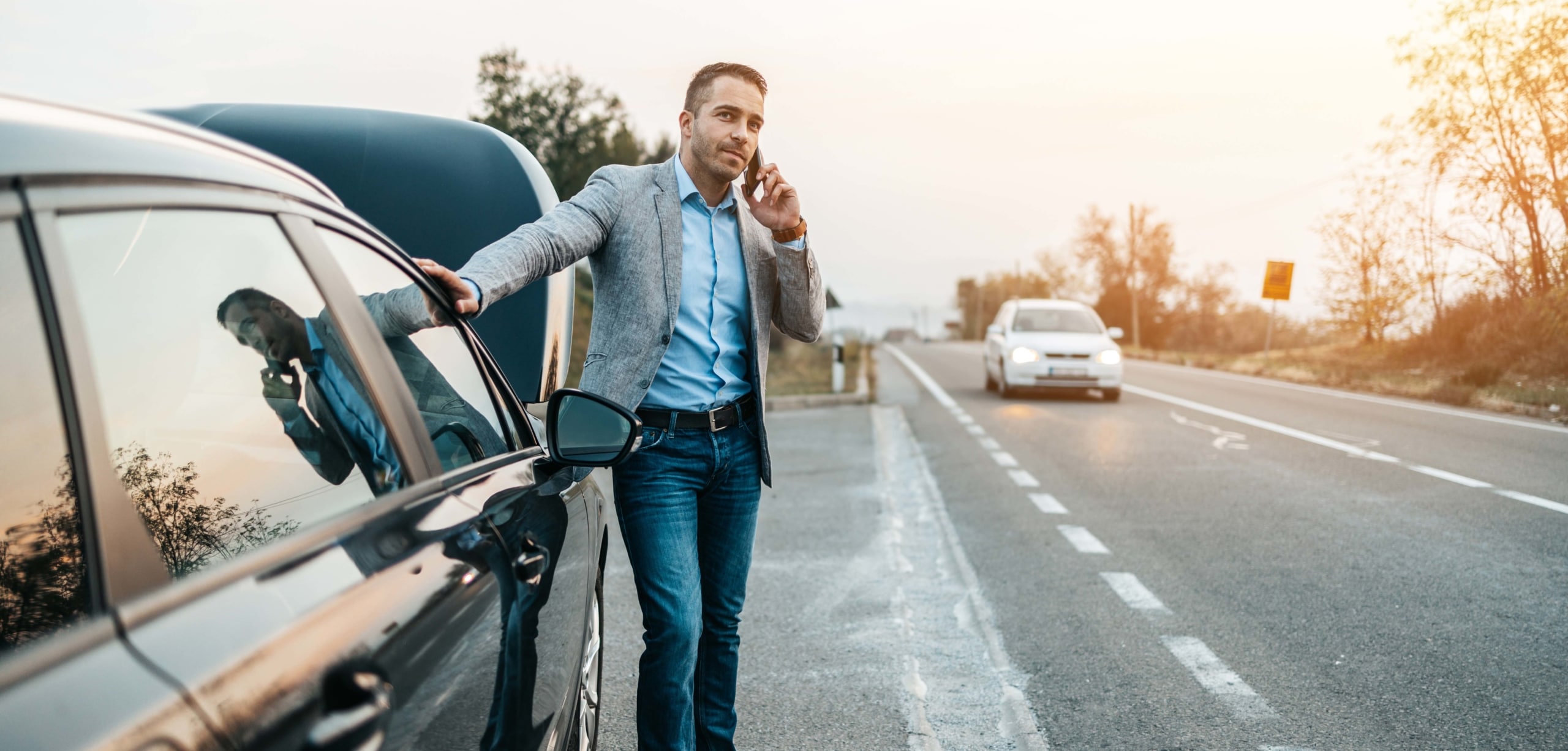 A middle-aged man in a blazer stands next to his car with a popped hood calling roadside assistance | Your Guide to Roadside Assistance Plans