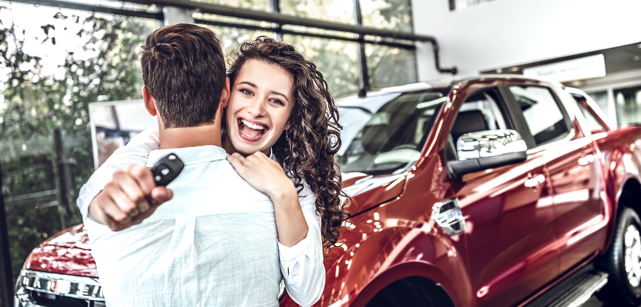 Excited woman is hugging a man and holding a set of car keys toward to camera with a new red truck in the background | Best Time to Save Big When Buying a Car