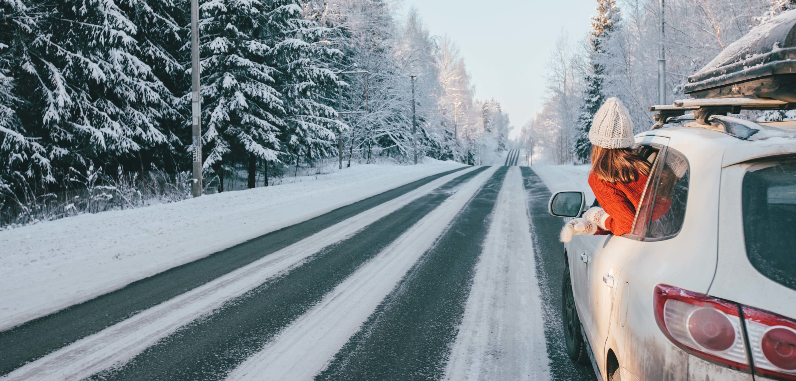 A woman in a knit hat sticks her head out the window on a scenic snowy highway | Get Your Car Holiday Road Trip Ready