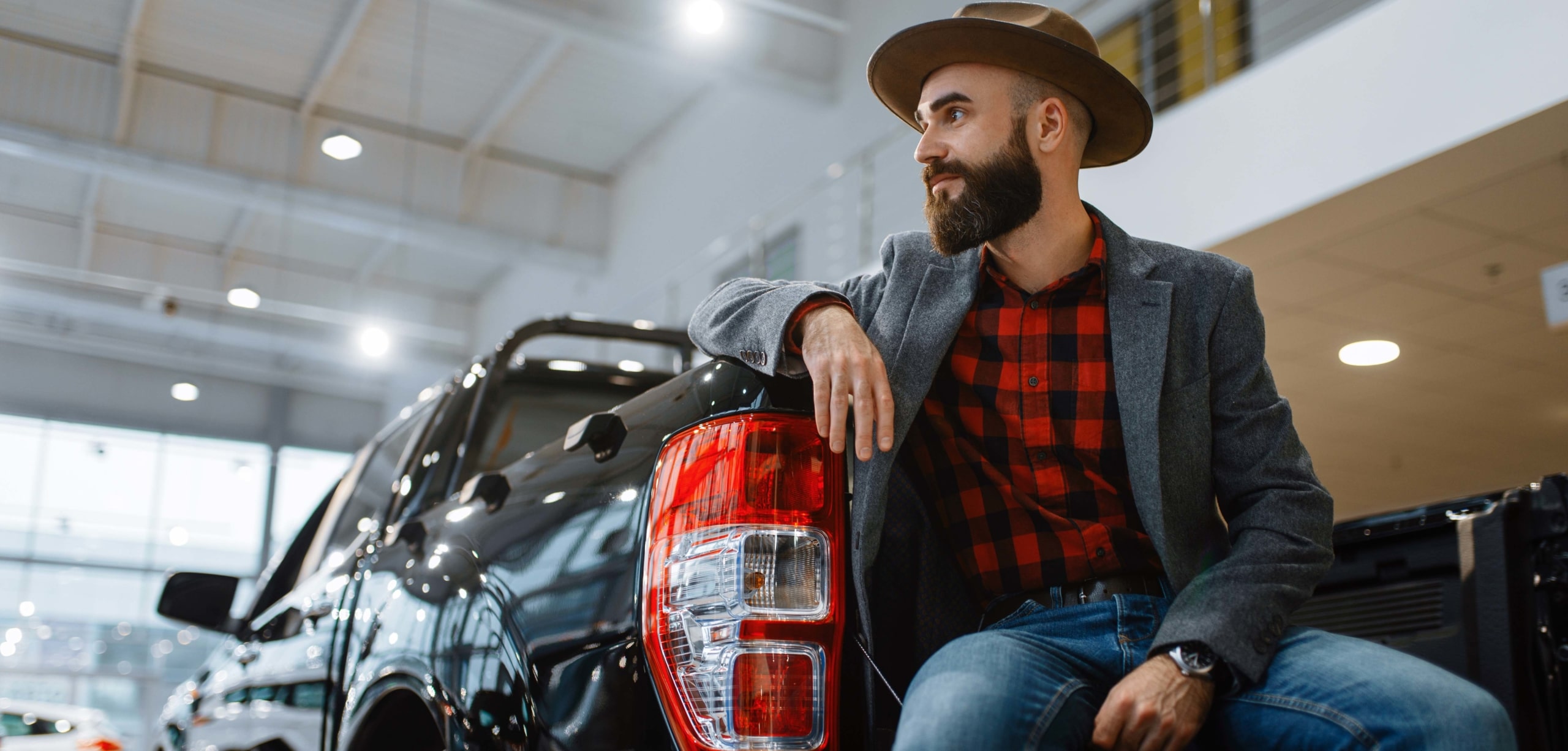 Man with a beard wearing a plaid shirt and a hat sits on the tailgate of a black truck at the dealership | Should I Buy a Car or Truck?