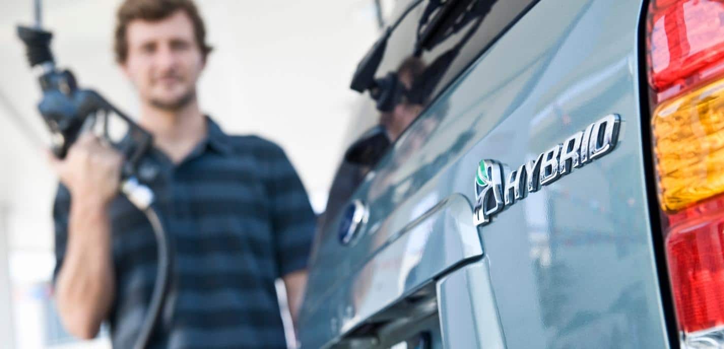 Close-up of a hybrid emblem on a car with a man holding a gas pump in the background