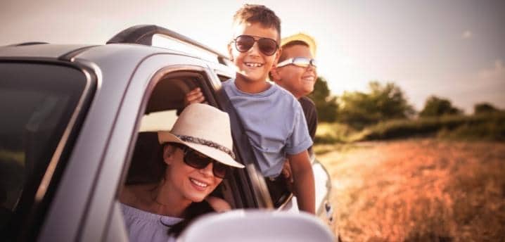 Family on a summer road trip with two young boys leaning outside of a car window while their mom is in the front seat