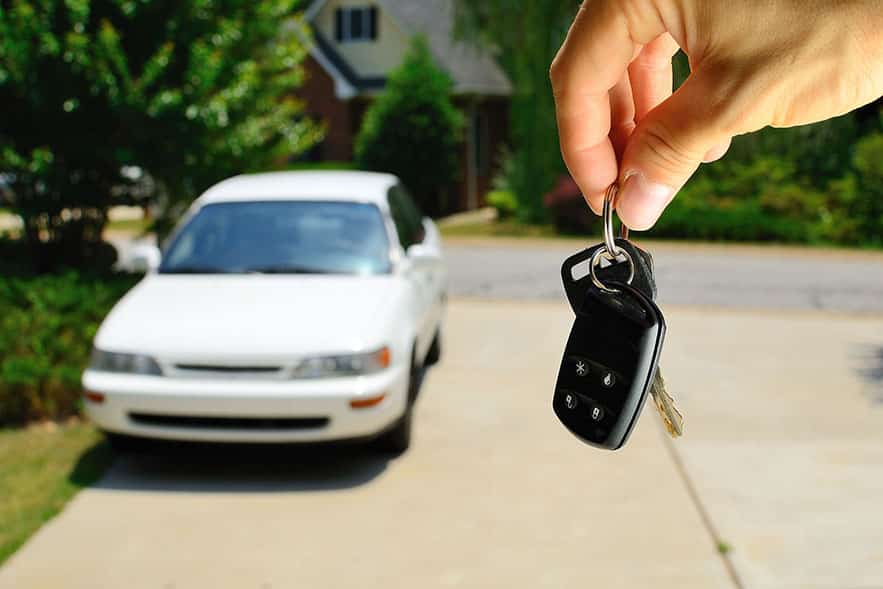 Person dangling a set of keys in the foreground with a car parked in the driveway in the background