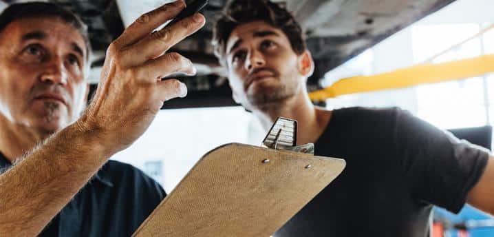 Two mechanics looking at the underside of a car while filling out a report