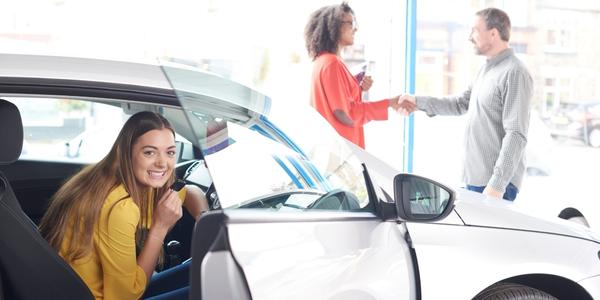 Teenager sitting in a new car with her parent in the background shaking hands with the salesperson