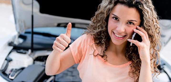 woman giving a thumbs up while on a cell phone, standing in front of a car