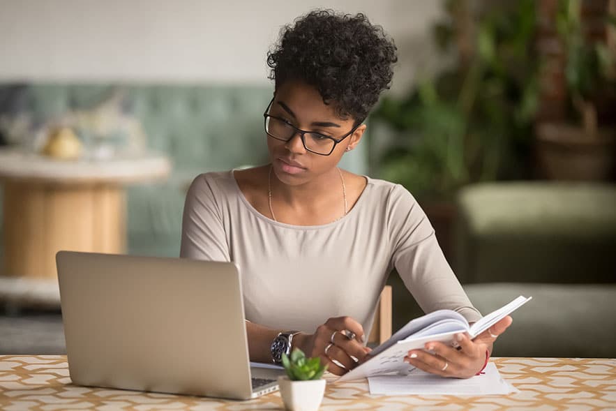 Woman looking at a computer screen with a notebook in her other hand as she makes notes