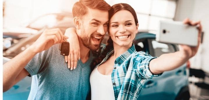 Young couple buying a car and taking a selfie with the keys and car in the background